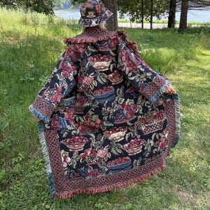 Könnte beinhalten: Ein schwarzer Wandteppichmantel und passender Fischerhut mit Blumen- und Fruchtdesign. Der Mantel hat einen gefransten Saum und Ärmel. Der Hut hat ein Kaffeetassen-Design. Der Mantel hat die Worte "Old Salem" am Saum.