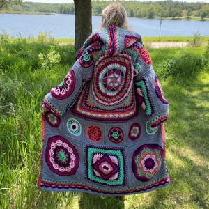 May include: A long, hooded crochet coat in shades of grey, pink, green, orange, and purple. The coat features a variety of geometric and floral patterns. The person wearing the coat is standing in front of a lake.