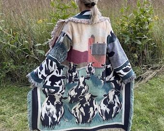 May include: A long, open-front woven cardigan featuring a pastoral scene with black and white cows grazing in a green field, a red barn, and a silo. The garment has a fringed hem and sleeves, with a repeating cow print pattern on the lower sections.