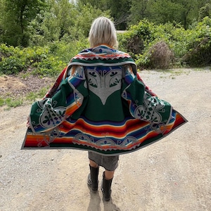 Könnte beinhalten: Ein farbenfrohes Poncho mit Muster, mit einem grünen Grund und einem grauen zentralen Design. Der Poncho hat ein Regenbogenstreifenmuster am unteren Rand und komplizierte geometrische Muster an den Rändern. Die Person trägt einen Jeansrock und schwarze Stiefel.
