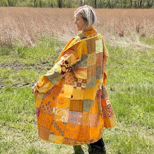 May include: A patchwork coat in shades of yellow, orange, and brown. The coat features a variety of patterned fabric squares sewn together. The long coat is worn outdoors in a field of green grass and tall brown grass.