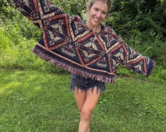 May include: A long-sleeved, cropped pullover top featuring a woven tapestry-style fabric with a dark, multi-colored geometric pattern in shades of black, rust orange, and cream, finished with a frayed fringe hemline along the bottom edge.