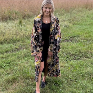 May include: A long-sleeved, open-front duster coat featuring a black base with a multi-colored floral and geometric pattern, layered over a black tank dress and paired with black loafers. The garment has visible white stitching throughout the fabric.