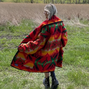 Op de afbeelding: Een persoon draagt een lange jas met een patroon met een levendig ontwerp in rood, geel, groen en zwart. De jas heeft een brede kraag en wordt over een donkere outfit gedragen. De persoon staat in een grasveld.