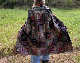 May include: A long coat constructed from a patchwork of various fabric squares in muted tones of brown, blue, green, and red, featuring a log cabin quilt pattern throughout the garment, paired with light blue wide-leg denim trousers.