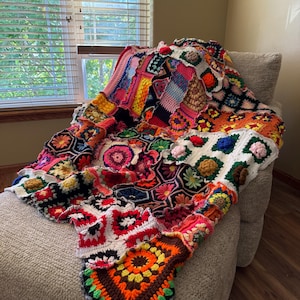 May include: A colorful, handmade crochet blanket draped over a beige armchair. The blanket features a patchwork design with various geometric shapes and floral patterns in shades of red, orange, yellow, green, and blue. The blanket is approximately 60 inches wide and 70 inches long.