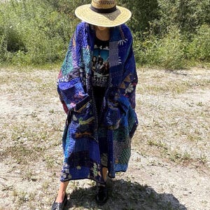 May include: A long, open-front coat made from blue patchwork fabric with visible kantha-style stitching. The coat features various shades of blue, patterned blocks, and deep pockets. It is worn over a black graphic t-shirt and paired with a straw hat.