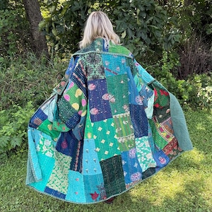 May include: A long, open-front jacket constructed from a patchwork of quilted fabric squares in various shades of green, blue, and teal. The garment features diverse floral, paisley, and geometric patterns with visible running stitch detailing.