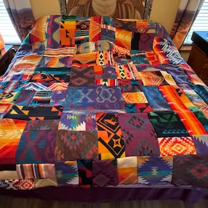 Könnte beinhalten: Eine Patchwork-Decke bestehend aus einer Vielzahl rechteckiger Stoffbahnen mit unterschiedlichen geometrischen Mustern und leuchtenden Farben wie Orange, Violett, Blau, Rot und Gelb. Die Decke liegt auf einem Bett und zeigt eine komplexe Anordnung gewebter Designs und kontrastierender Farbtöne über die gesamte Fläche.