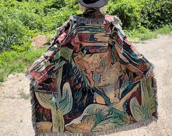 May include: A long, open-front tapestry coat featuring a desert landscape print with cacti, rock formations, and a starry night sky in shades of teal, red, and tan. The garment has frayed edges along the hem and sleeves, paired with a black cowboy hat.