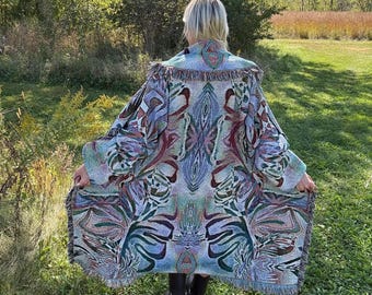 May include: A colorful, patterned woven coat with fringe trim. The coat features a symmetrical design in shades of blue, green, purple, and cream. The person is standing in a grassy field with trees in the background.