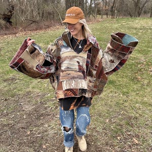 May include: A patchwork jacket with a landscape scene, featuring a pheasant, is worn over a black shirt and distressed jeans. A brown cap with white text is worn. The jacket has brown, tan, and burgundy patches.