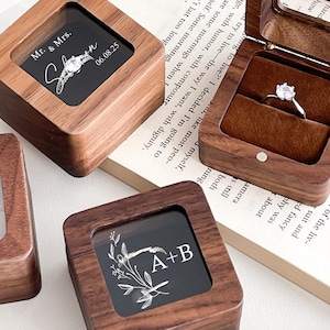 Pode incluir: Caixas de anel de madeira com tampas transparentes. Algumas caixas estão abertas, revelando um anel e um interior de veludo. Outras apresentam iniciais e datas gravadas. As caixas são de cor castanho escuro.
