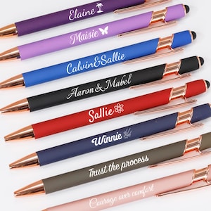 Op de afbeelding: Een assortiment van acht pennen in verschillende kleuren, waaronder paars, lavendel, blauw, zwart, rood, marine, olijf en roze. Elke pen heeft een roségouden accent en een naam of zin op de schacht. De pennen liggen op een witte ondergrond.