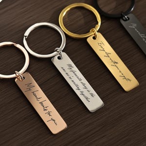 Peut inclure: Quatre porte-clés gravés en or rose, argent, or et noir. Chaque étiquette rectangulaire porte une inscription différente en écriture cursive. Les porte-clés sont sur une surface en bois brun foncé.