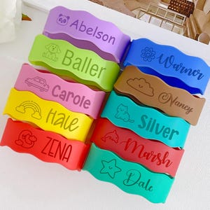 May include: A collection of colorful, wavy silicone wristbands. Each band features a different name and a small graphic, such as a car, cloud, or star. The bands are in various colors, including purple, green, blue, brown, pink, yellow, red, and teal.
