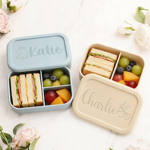May include: Two personalized lunch boxes, one light blue and one beige, each with a name and a cartoon animal. The blue box says "Katie" with a unicorn, and the beige box says "Charlie" with a llama. Both boxes are open, revealing sandwiches and fruit.
