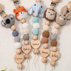 May include: A collection of baby pacifier clips with wooden beads and animal-shaped crochet toppers. Each clip features a different animal: a koala, fox, elephant, lion, and bunny. Personalized wooden heart-shaped charms are engraved with names.