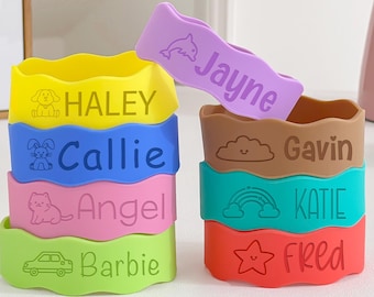May include: A stack of colorful silicone bands with names and small graphics. The bands are yellow, blue, pink, green, brown, teal, and red. Each band has a different name, such as "Haley," "Callie," and "Fred." A purple band with the name "Jayne" is on top.
