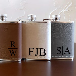 May include: Three stainless steel hip flasks with leatherette coverings. The flasks are brown, beige, and gray. The flasks are engraved with the initials "R/W", "FJB", and "S/A".