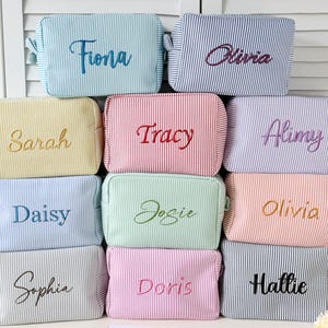 Può includere: Una collezione di trousse personalizzate in vari colori, tra cui blu, giallo, rosa e verde. Ogni trousse presenta un motivo a righe verticali e un nome ricamato in un colore complementare. I nomi includono Fiona, Sarah, Daisy, Sophie, Olivia, Tracy, Alimy, Josie, Olivia, Doris e Hallie.