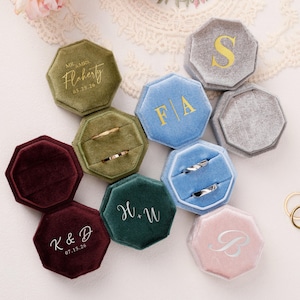 Può includere: Un assortimento di scatole per anelli in velluto di vari colori, tra cui verde, blu, bordeaux e rosa. Alcune scatole sono personalizzate con iniziali, nomi e date in lettere dorate o argentate. Sono visibili fedi nuziali d'oro.