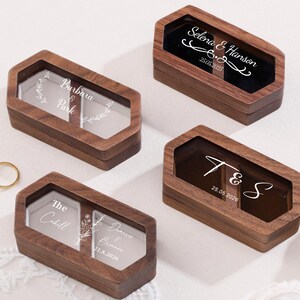 May include: Four wooden ring boxes with clear tops, each engraved with names and dates. The boxes are a dark brown color and have a hexagonal shape. Two gold wedding rings are visible in the image. The boxes are likely for wedding rings.