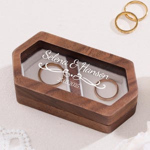Puede incluir: Un joyero de madera con tapa transparente, que contiene dos alianzas de oro. La caja está grabada con los nombres "Selena & Hanson" y la fecha "25.05.2025". Dos anillos de oro adicionales son visibles en el fondo.
