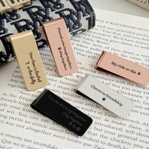 May include: A collection of metal money clips in gold, rose gold, black, and silver. Each clip has an engraved message, including "Your time is limited I so live fully" and "Friends who laugh together stay together."