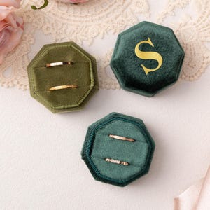 May include: Three velvet ring boxes in shades of green, each with a hexagonal shape. Two boxes hold gold rings. One box has a gold letter "S". The boxes are on a white surface with floral and lace accents.