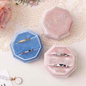 May include: Three velvet ring boxes in blue and pink, each holding rings. The pink box has "K & D 07.13.26" printed on the lid. The rings are gold and silver, with various designs. The boxes are octagonal.