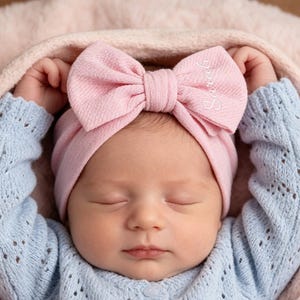 Op de afbeelding: Een pasgeboren baby met een lichtblauwe trui en een roze haarband met strik en de naam "Sarah" geborduurd. De baby is gewikkeld in een roze deken en heeft de ogen gesloten.