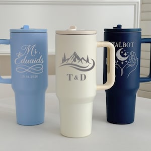 May include: Three insulated travel mugs with handles and lids. One is light blue with "Mr. Eduaids" and a date. The second is cream-colored with a mountain graphic and "T&D". The third is navy blue with "Talbot" and a celestial design.