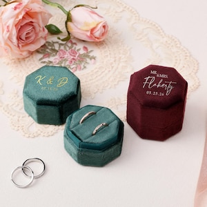 May include: Three velvet ring boxes in teal and burgundy. One teal box is open, displaying two rings. The boxes are octagonal and have gold lettering. The burgundy box has the text "Mr. & Mrs. Flaherty" and a date. Pink roses and lace are in the background.