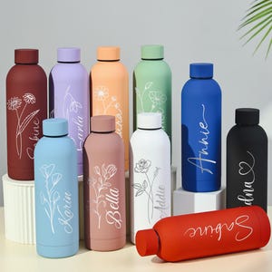 Peut inclure: Une collection de bouteilles d'eau personnalisées colorées. Chaque bouteille, de couleurs variées comme le rouge, le bleu et le vert, porte un nom et un motif floral en écriture blanche. Les bouteilles sont cylindriques avec un bouchon à vis.
