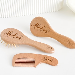 May include: A wooden baby grooming set comprising a brush, a rounded brush, and a comb. Each item is engraved with the name "Abadam" in a script font. The brush has soft bristles. The wood has a light, natural finish. The comb measures approximately 10 cm.