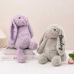 May include: Two plush bunny rabbits, one lavender and one gray, with pink noses and embroidered names. The lavender bunny has "Gabriel" embroidered on its ear, and the gray bunny has "Dailey" embroidered on its ear. The bunnies are sitting on a stack of books.