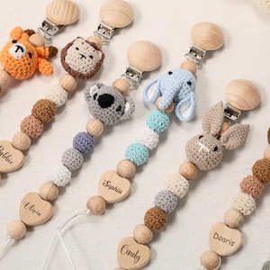 Może przedstawiać: Kolekcja klipsów do smoczków dla niemowląt z drewnianymi koralikami i szydełkowymi wzorami zwierząt. Każdy klips przedstawia inne zwierzę, w tym lwa, koalę, słonia i królika. Klipsy mają drewniane serce z wygrawerowanym imieniem.