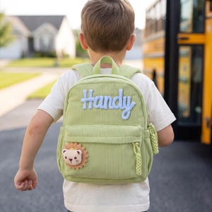 May include: A light green corduroy backpack featuring the word Handy in light blue script lettering across the top. The bag includes a small lion-shaped patch on the lower left corner and braided zipper pulls on the front pocket and main compartment.