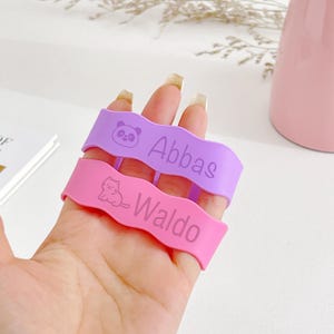 May include: Two silicone wristbands, one lavender and one pink, are held up. The lavender band has a panda graphic and the name "Abbas." The pink band has a cat graphic and the name "Waldo."