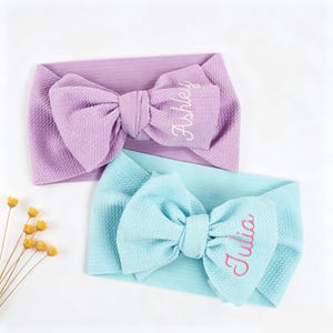 May include: Two baby headbands, one lavender and one light blue, each with a large bow. The lavender headband has the name "Ashley" embroidered in white, while the blue headband has "Julia" embroidered in pink.