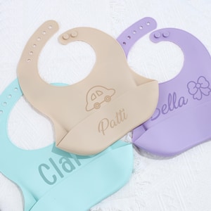 May include: Three silicone baby bibs in beige, light blue, and purple. The beige bib has a car graphic and the name "Patti". The purple bib has a bow graphic and the name "Bella". The light blue bib has the name "Cla".