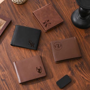 May include: Four leather wallets in black and brown are displayed on a wooden surface. Each wallet is personalized with initials, names, or a graphic. The wallets are closed and rectangular, showcasing a classic design.