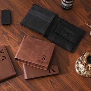May include: An open black leather wallet with a "Happy birthday, I love you - Emily" inscription is displayed next to two brown leather wallets. One brown wallet is embossed with the letter "S" and the name "Samuel". A black smartphone is also visible.