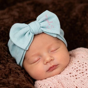 Pode incluir: Um bebê dormindo usando uma faixa de cabelo com laço azul claro bordada com o nome "Sophia" em rosa. O bebê está usando um suéter rosa e descansando em uma superfície marrom texturizada.