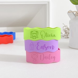 May include: A stack of three colorful silicone baby bottle bands. The top band is green and says "Olivia" with a girl icon. The middle band is purple and says "Eason" with a bottle icon. The bottom band is pink and says "Dailey" with a boy icon.