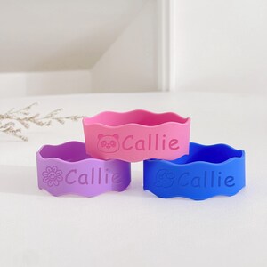 May include: Three colorful silicone cup sleeves in pink, purple, and blue. Each sleeve has a wavy top edge and the name "Callie" printed on it. The pink sleeve features a panda, the purple a flower, and the blue a cartoon character.