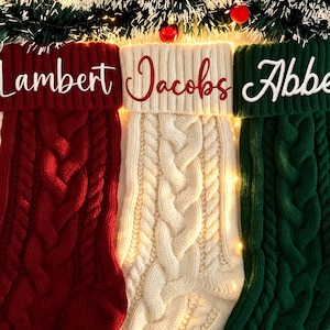 Puede incluir: Tres calcetines navideños personalizados en rojo, crema y verde, cada uno con un diseño de punto trenzado. Los nombres "Lambert", "Jacobs" y "Abbe" están bordados en los puños. Decoración festiva navideña.