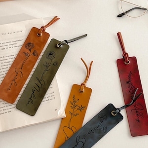 Könnte beinhalten: Lesezeichen aus Leder in verschiedenen Farben, darunter Braun, Grün, Gelb, Blau und Rot. Jedes Lesezeichen hat ein Blumendesign und einen Namen. Sie haben eine Lederschlaufe oben.