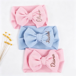 Op de afbeelding: Drie baby haarbanden in roze en blauw. Elke haarband heeft een grote strik met een geborduurde naam: Olivia, Rachel en Diana. De haarbanden zijn gemaakt van een zachte, getextureerde stof.